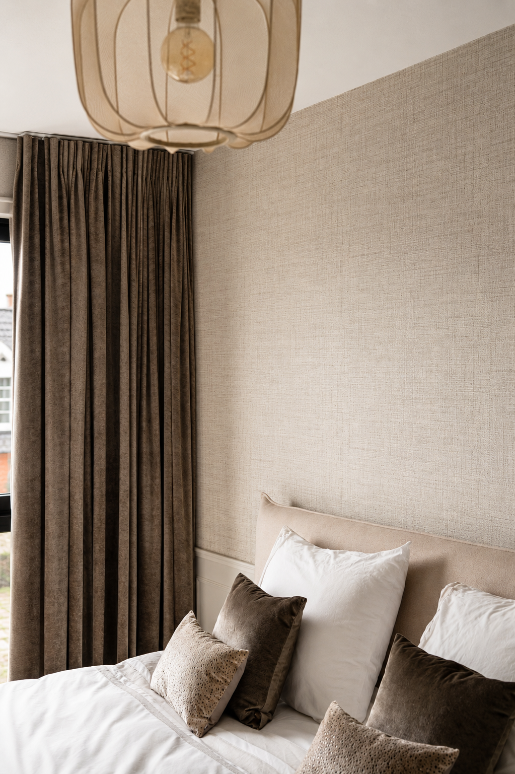 Gestoffeerd beige hoofdbord met decoratieve kussens en velours gordijnen in warme aardetinten.